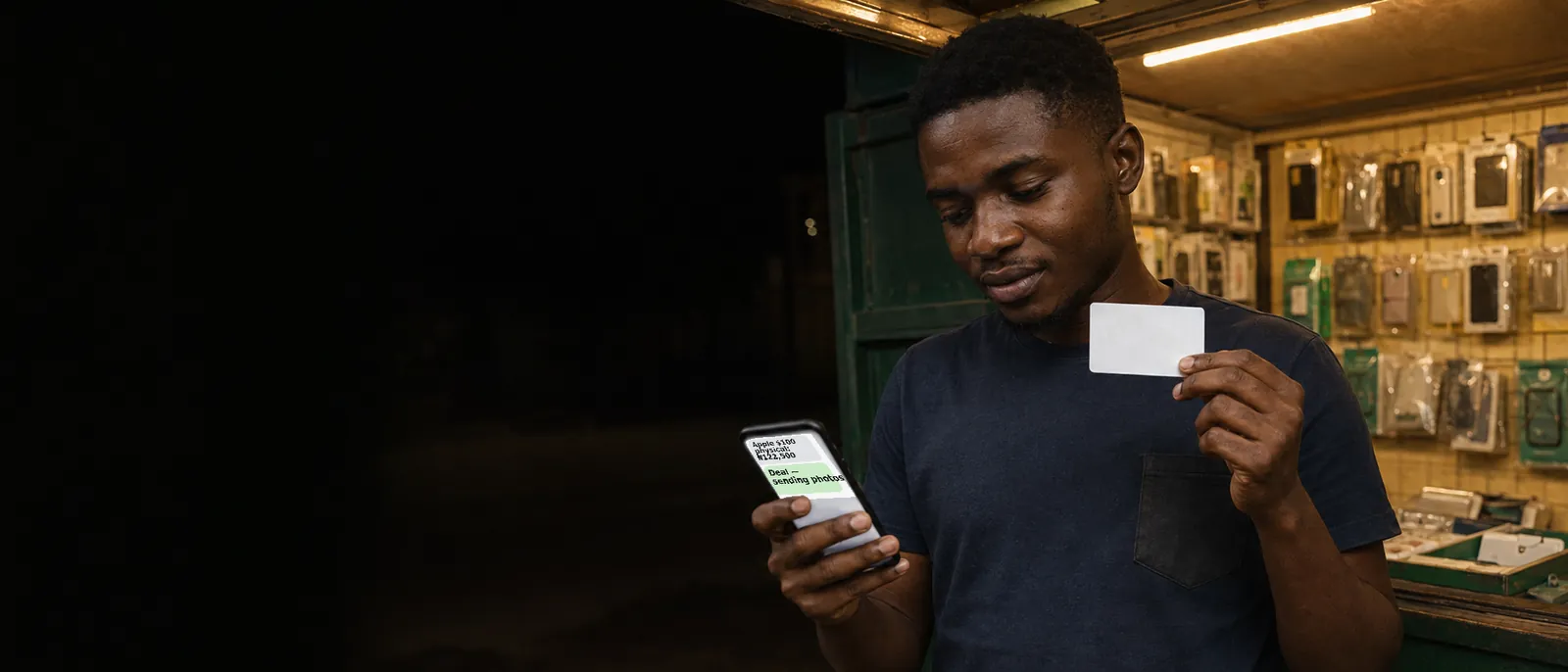 A young Nigerian man at a small phone-accessories kiosk at dusk, warm light on shelves of packaged accessories behind him, holding a blank white gift card in one hand and a phone showing a chat quoting ₦122,500 for a $100 Apple card in the other