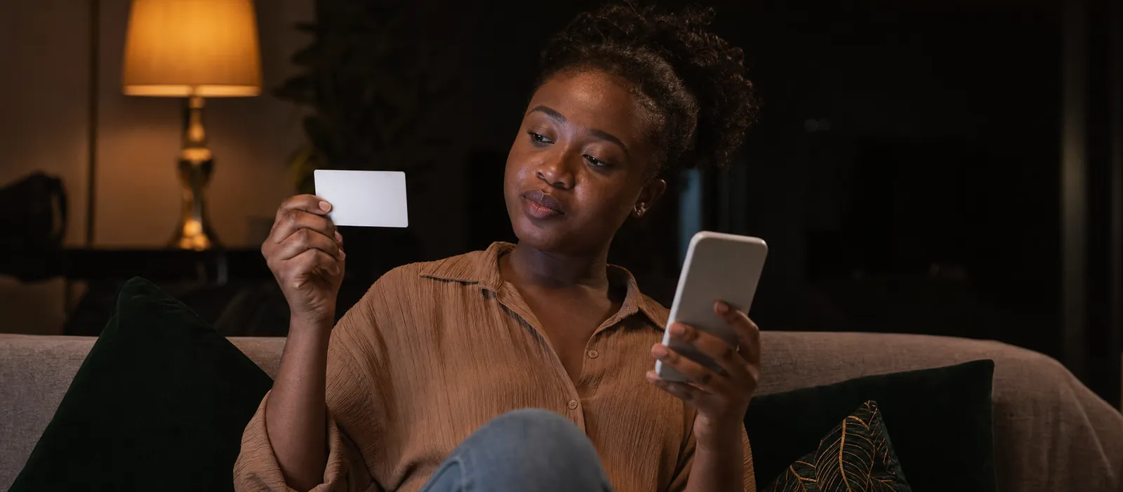 A young Nigerian woman on a living-room sofa in warm evening lamplight, holding a blank white gift card up beside her iPhone and comparing the two