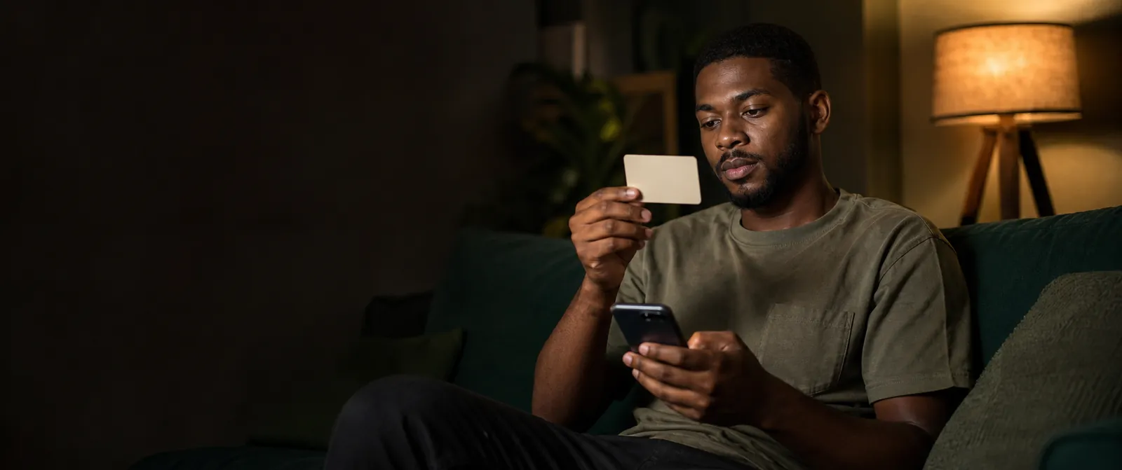 A young Nigerian man on a sofa in warm evening lamplight, holding up a gift card and typing its number into his phone