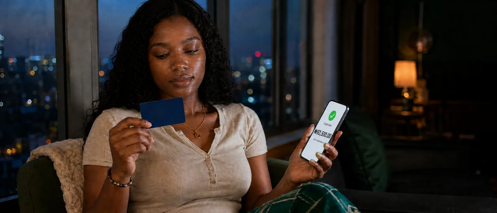 A young Nigerian woman by a large apartment window at dusk, city lights blurred outside, holding a blank dark-blue gift card in one hand and a phone showing a ₦93,500 credit alert in the other