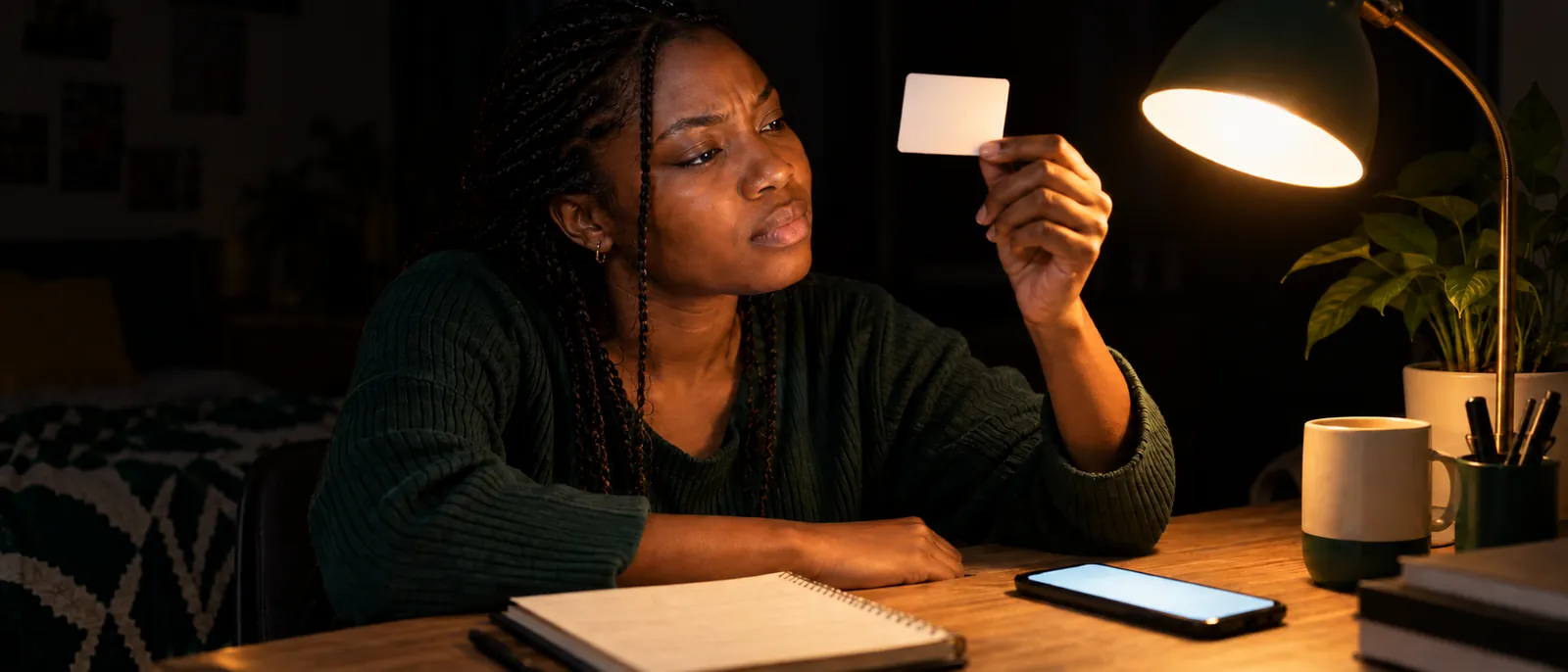 A young Nigerian woman at a home desk in the late evening, holding a plain matte-white prepaid card up toward a warm desk lamp as if reading its fine print, her phone glowing softly beside a notebook