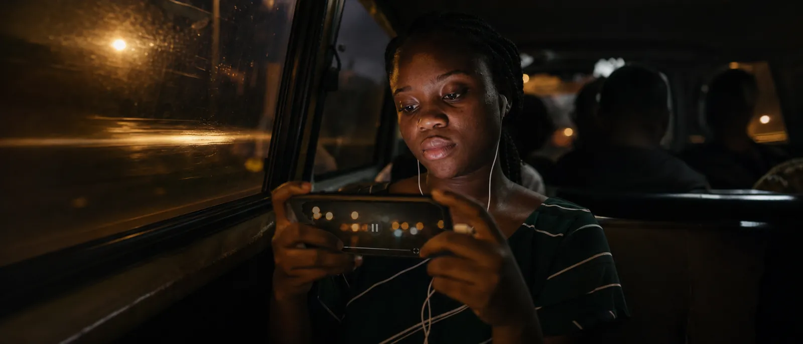 A young Nigerian woman in a danfo bus at dusk watching a video on a horizontally held smartphone, her face softly lit by the screen, warm street-light streaks across the window